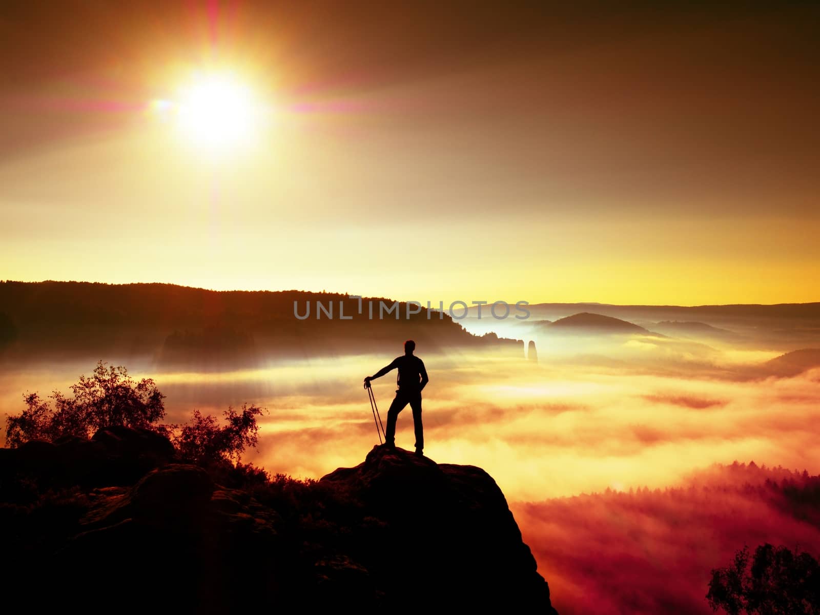 Tall backpacker with poles in hand. Sunny misty daybreak in rocky mountains. Hiker with backpack stand on rocky view point above valley. Vivid and strong vignetting effect.