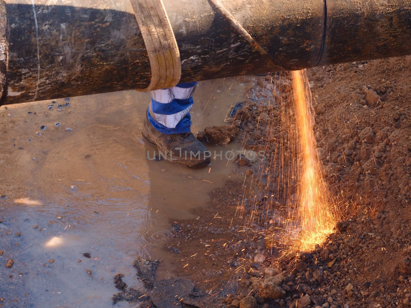 Worker staff is cutting big metal tube with hand grinder. Burning parks are flying down to wet clay. Staff use safety clothes, protective boots. Deep excavation pit.