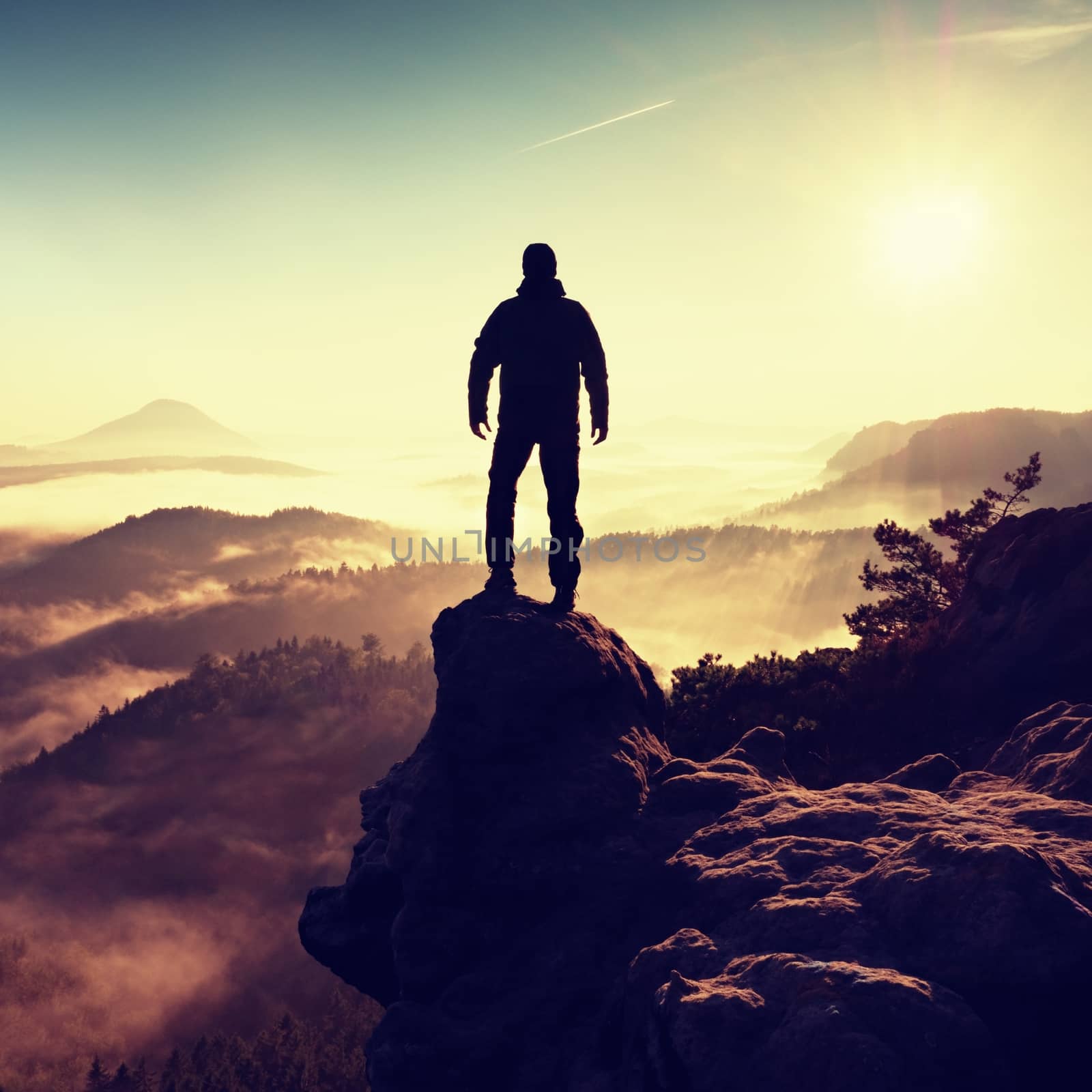 Man silhouette climbing high on cliff. Hiker climbed up to peak enjoy view. Man  watch over misty and foggy morning valley in morning Sun. 