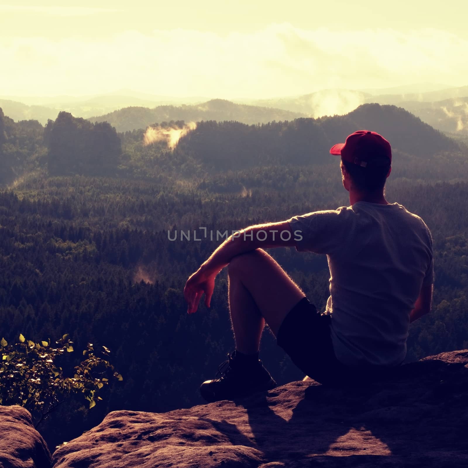 Man tourist sit on exposed rock. View point with sharp rock above misty valley. Sunny daybreak in rocky mountains.