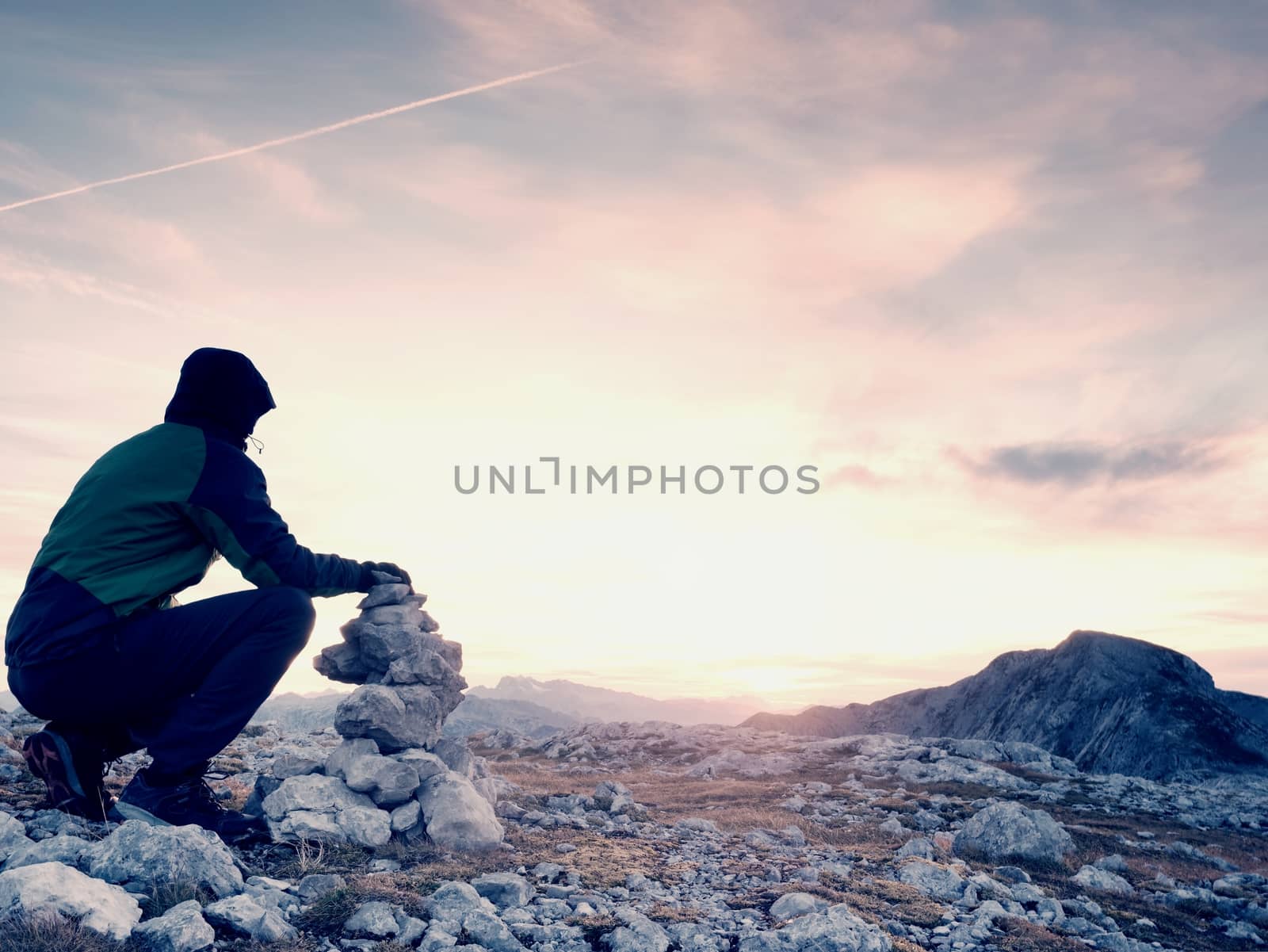Man hiker is building pebbles pyramid. Stones on Alps mountain summit. Daybreak horizon above blue foggy valley.