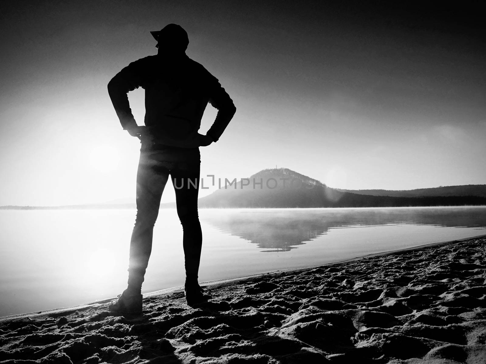 Man exercising on the beach.  Silhouette of active man exercising  and stretching on the lake beach at sunrise. Healthy lifestyle. Alone young fitness man exercise  at morning beach. Lens flare defect.