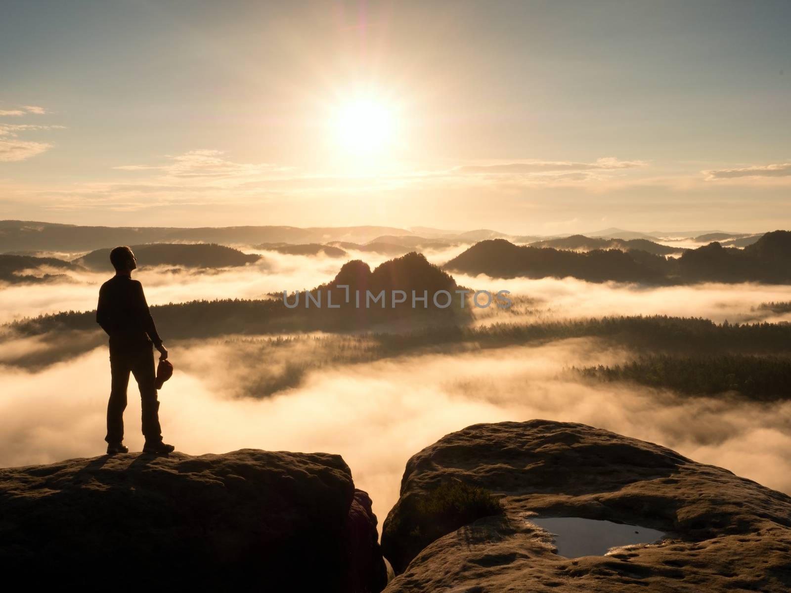 Hiker silhouette standing on rocky summit above misty valley. by rdonar2