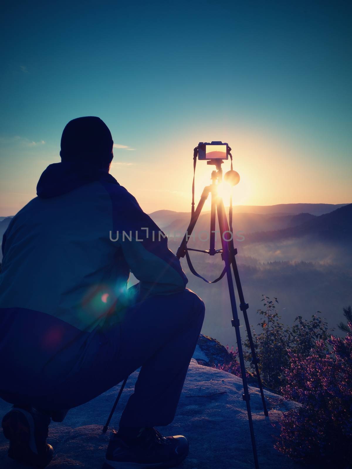 Nature photographer with tripod and camera on cliff and thinking. Dreamy fogy landscape, orange misty sunrise in a beautiful mountains. Vivid and strong vignetting effect.