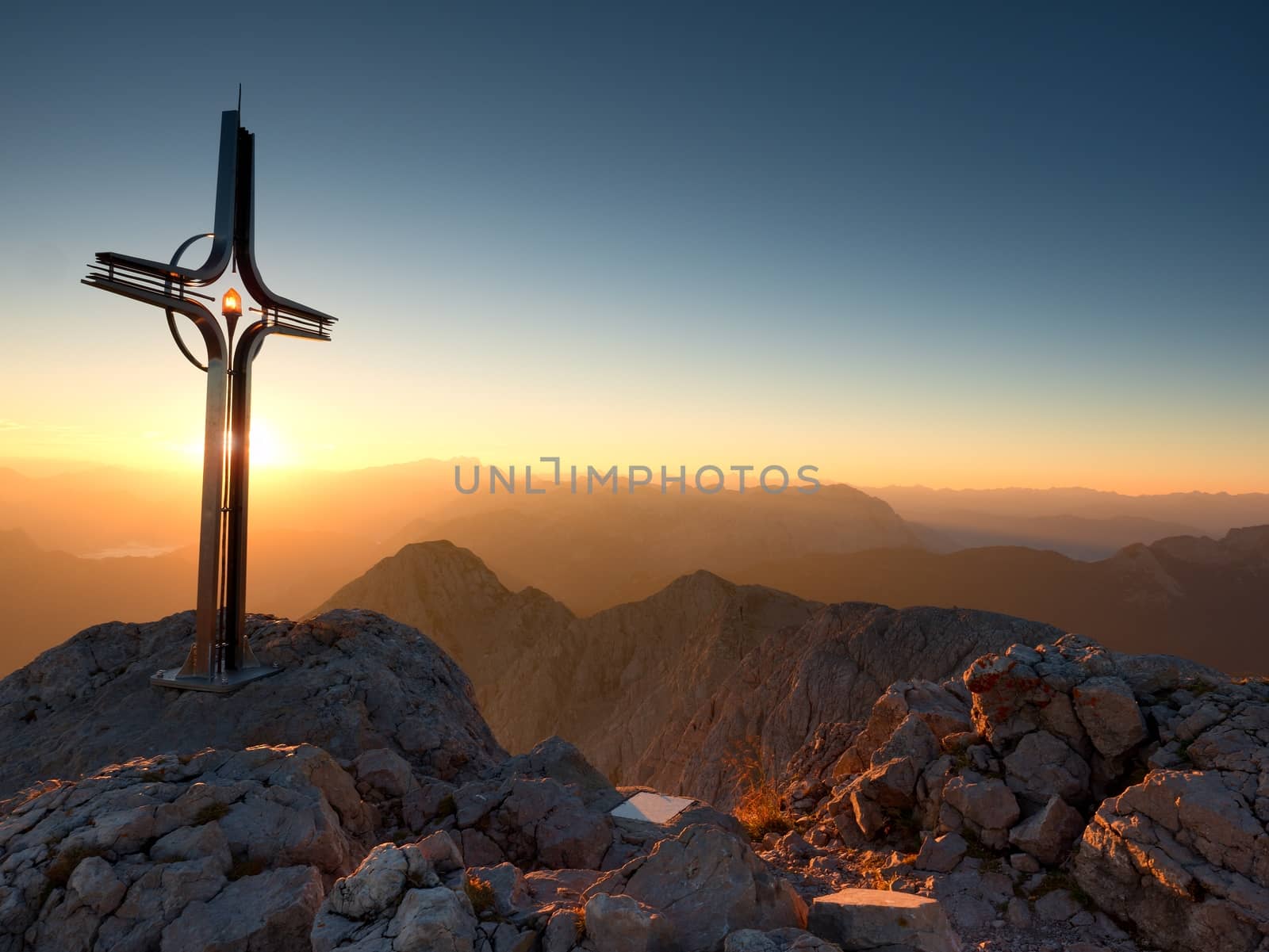 Cross raised  at mountain summit  in Alps. Sharp peak, daybreak Sun in sky. Steel crucifix in memory of victims of mountains. Vivid photo.