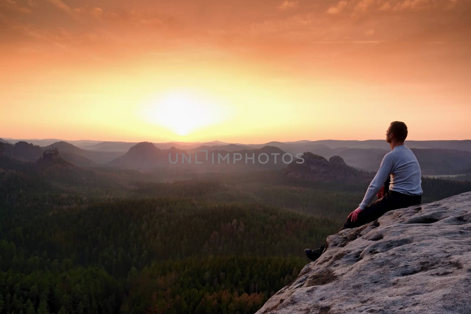 Man sit on the peak of rock and watching into colorful mist and fog in forest valley. Hiker enjoying view of morning nature