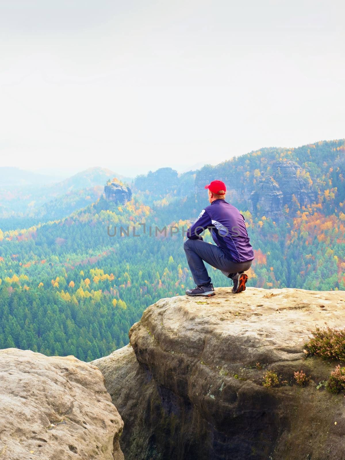 Moment of loneliness. Man in black enjoy marvelous view. Hiker sit on the peak of rock and watching into colorful mist and fog in forest valley.