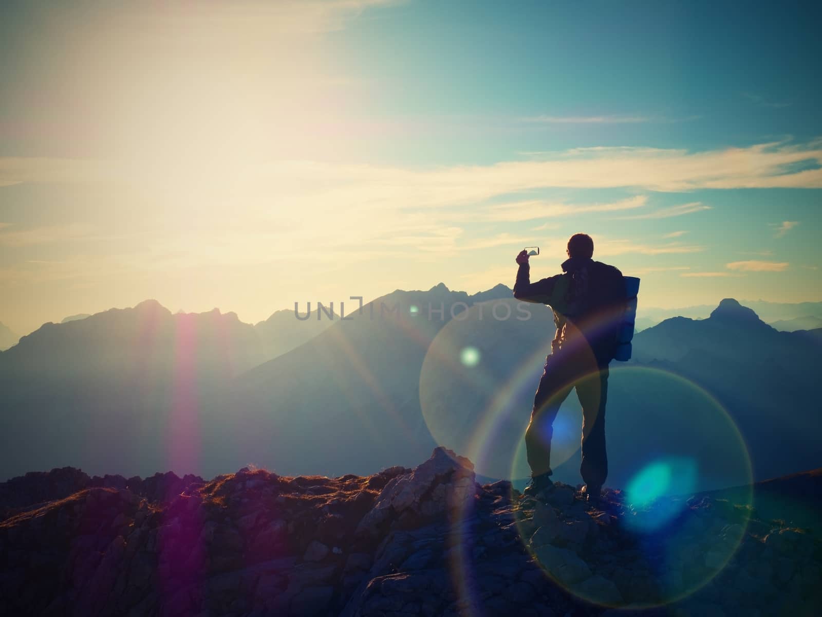 Lens flare light, strong defect. Hiker takes selfie photo. Man with big backpack and poles walk on Alpine mountain. Sun above horizon blue foggy valley. Mountains increased from humidity