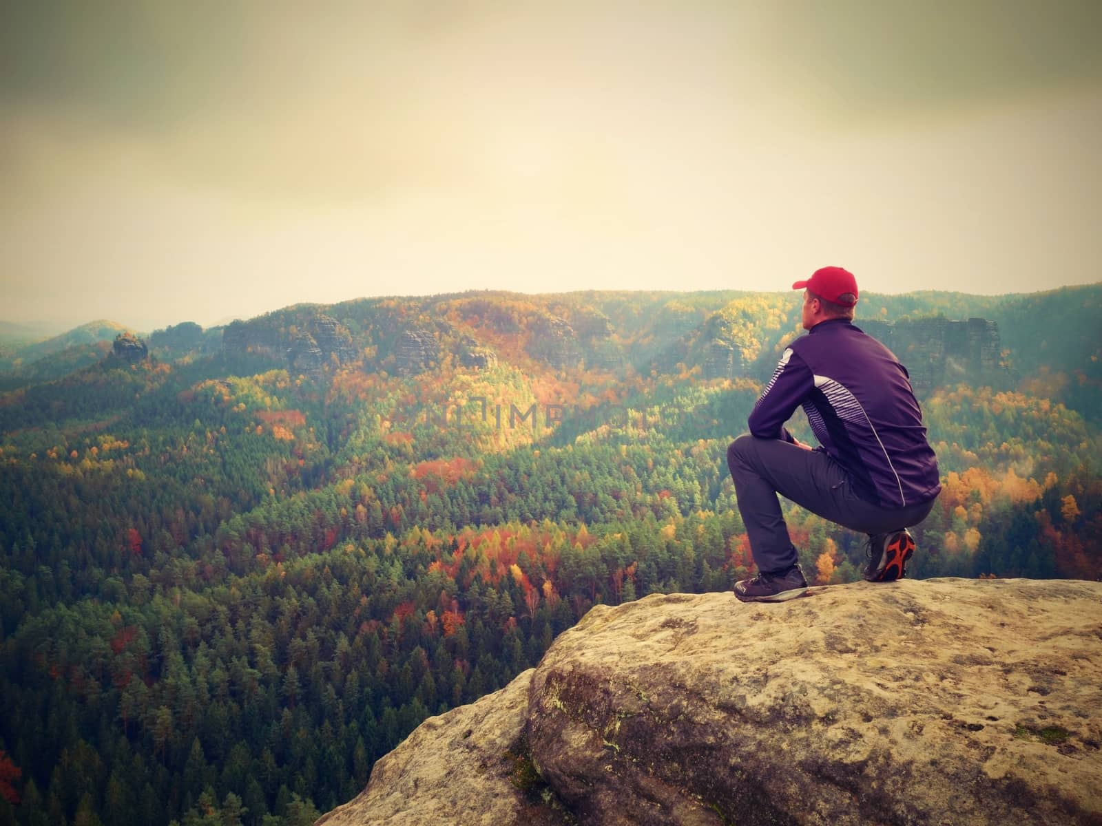Moment of loneliness. Man in black enjoy marvelous view. Hiker sit on the peak of rock and watching into colorful mist and fog in forest valley.