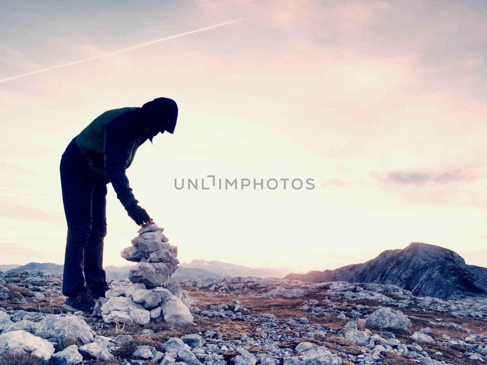 Alone adult man is stocking stone to pyramid.Alps mountain summit, evening sun at horizon.  Mountains increased from humidity
