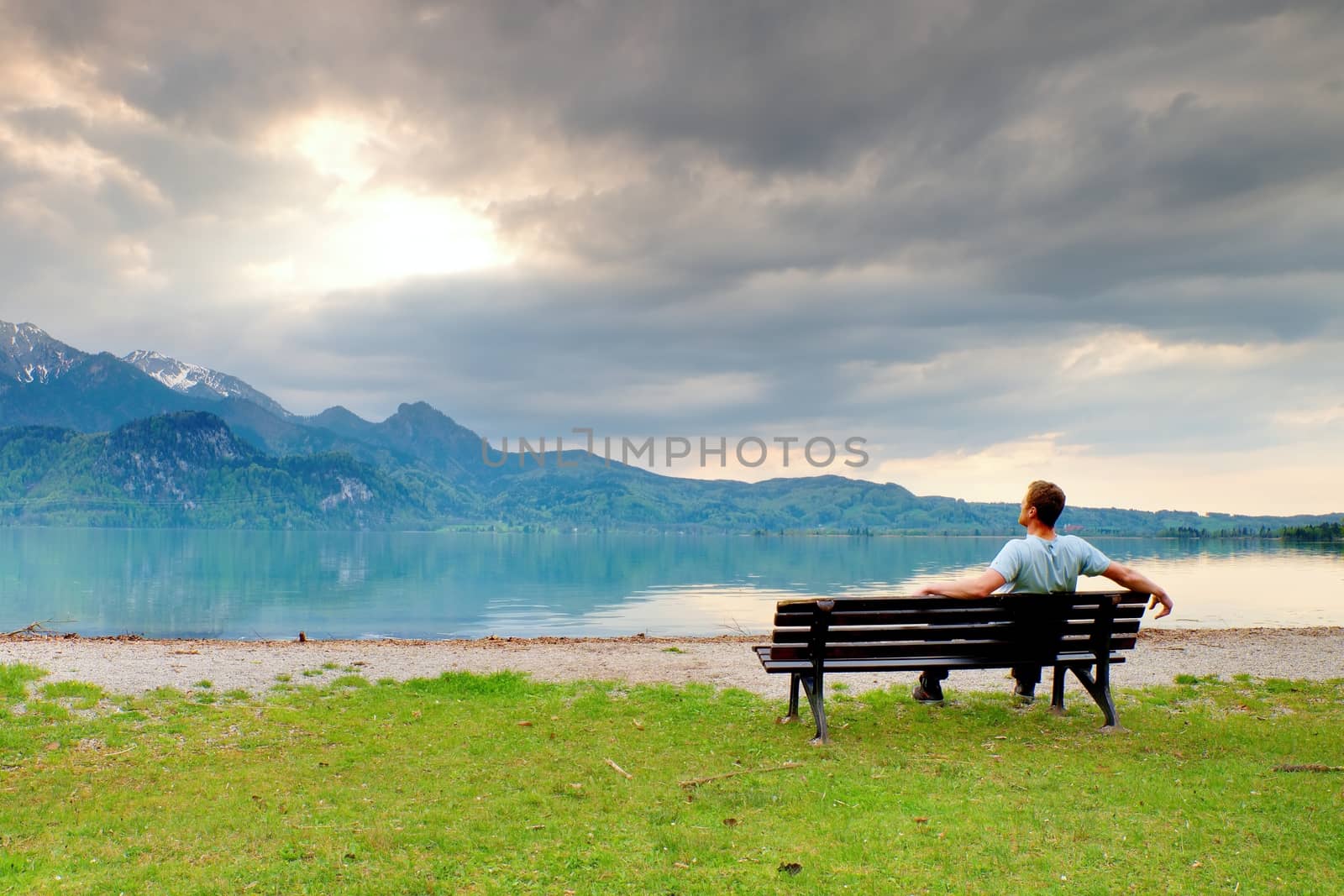 Alone  man sits on bench beside an azure mountain lake. Man relax and watch high peaks of Alps above lake mirror.