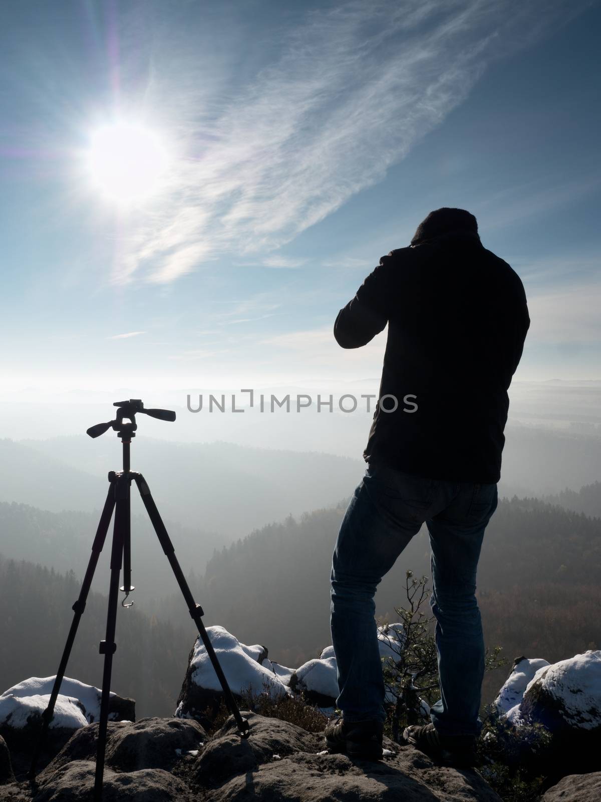 Nature photographer stay with camera ind hands  at tripod on rock and thinking. First snow. Dreamy fogy landscape, orange misty sunrise in  beautiful mountains