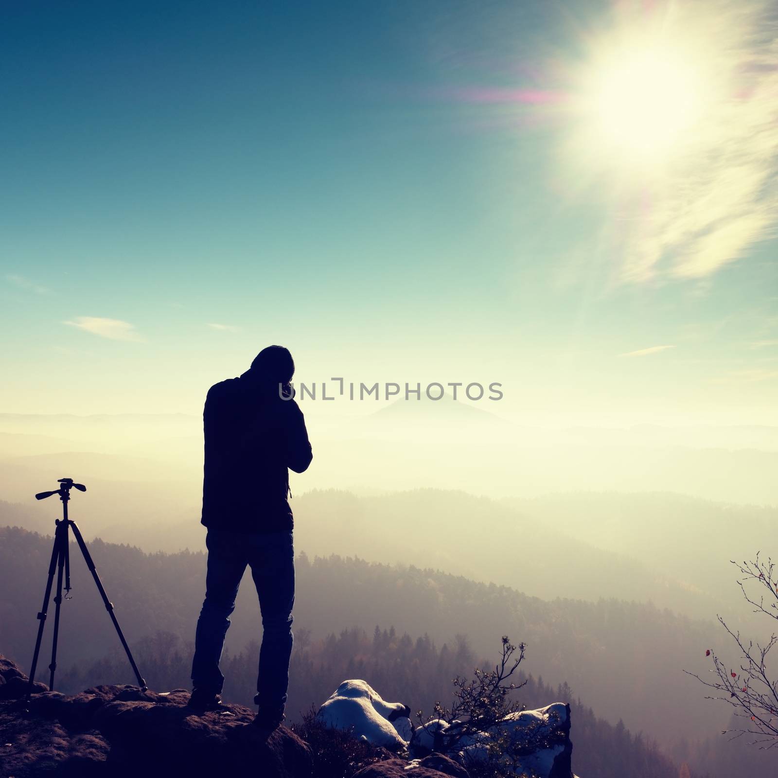 Professional nature photographer do work on snowy cliff.  Man takes photos with mirror camera on peak of rock. Dreamy fogy landscape, spring orange pink misty sunrise.