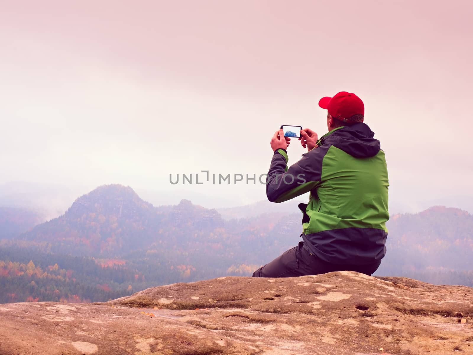 Man sitting on rock takes photos with smart phone. Hiker on peak of rock empire. Melancholy fogy landscape, spring orange pink misty sunrise in mountains.