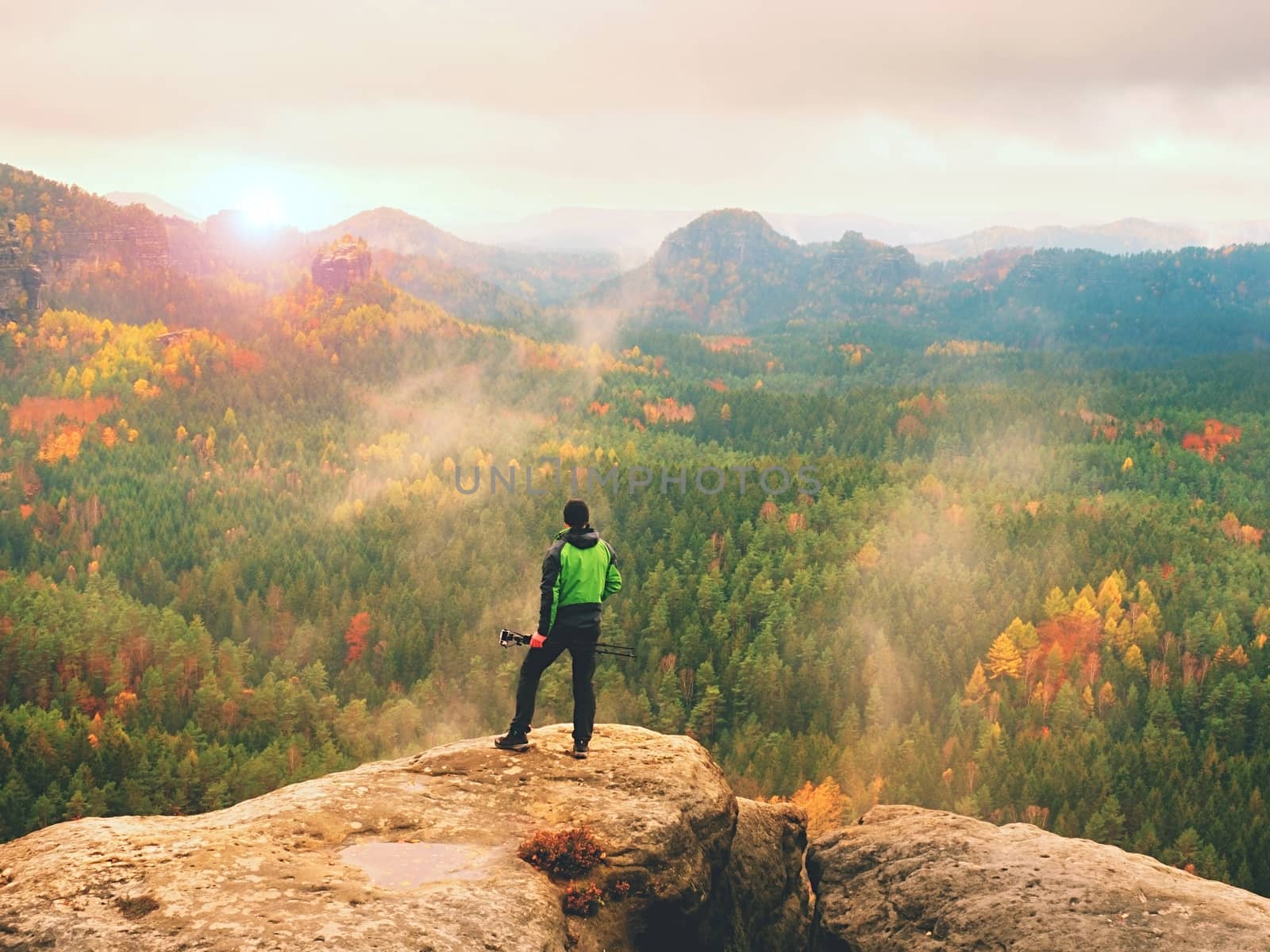 Tall adult  photographer prepare camera for taking picture of fall  mountains. Photograph at daybreak above  colorful  valley. Landscape view of autumn mountain hills and hiker above