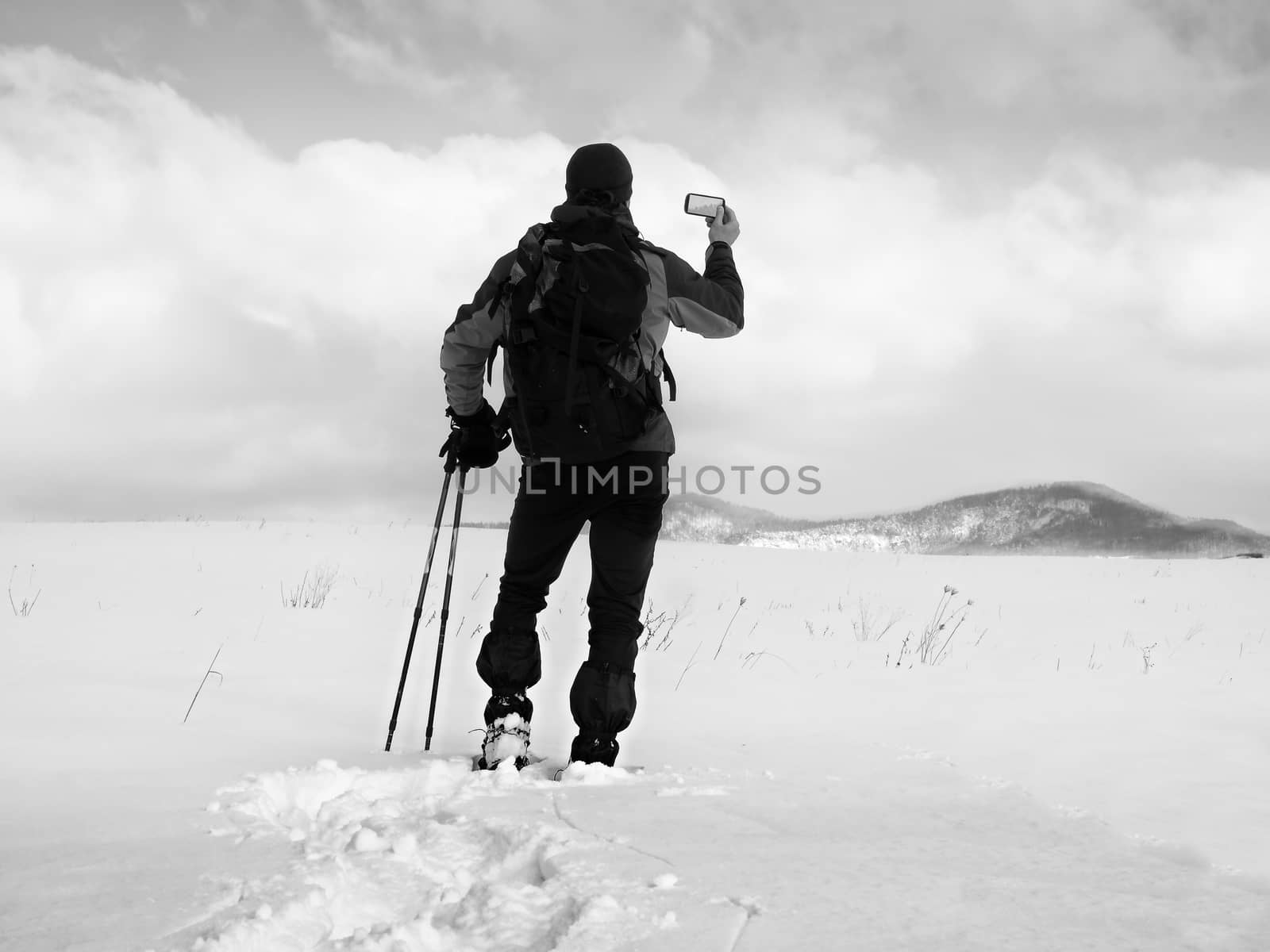 Man with snowshoes and backpack take photos by smartphone. Hiker in snowdrift,  snowshoeing in powder snow. Cloudy winter day, gentle wind brings small snow flakes 