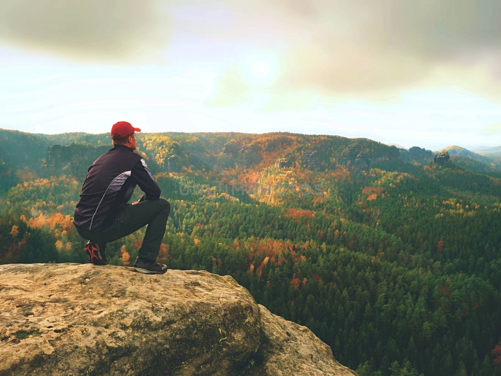 Man in black outdoor clothes sit on cracked rocky empires. Fall colorfull landcape. Melancholy misty day in sandstone mountains, mist in deep valley.