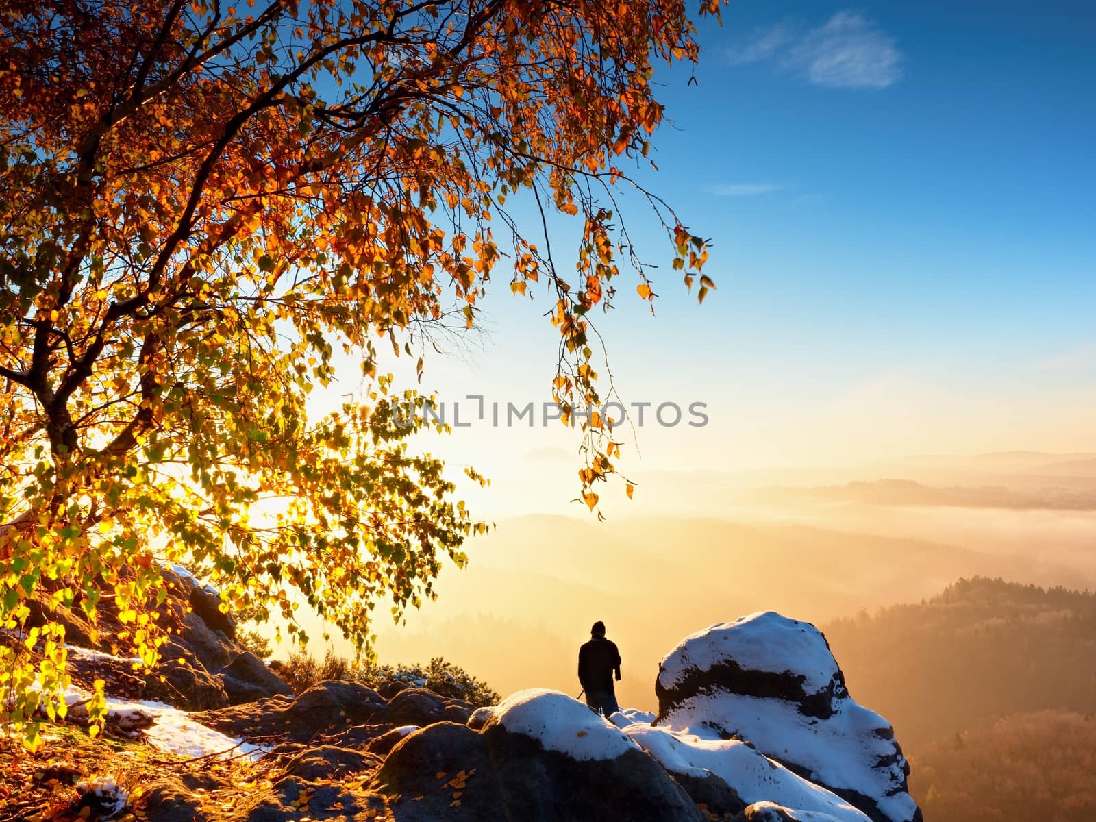 Sunny early winter  morning. Photographer preparing camera on tripod. Snowy rocks, in valley bellow colorful leaves forest. View over misty and foggy valley to Sun.