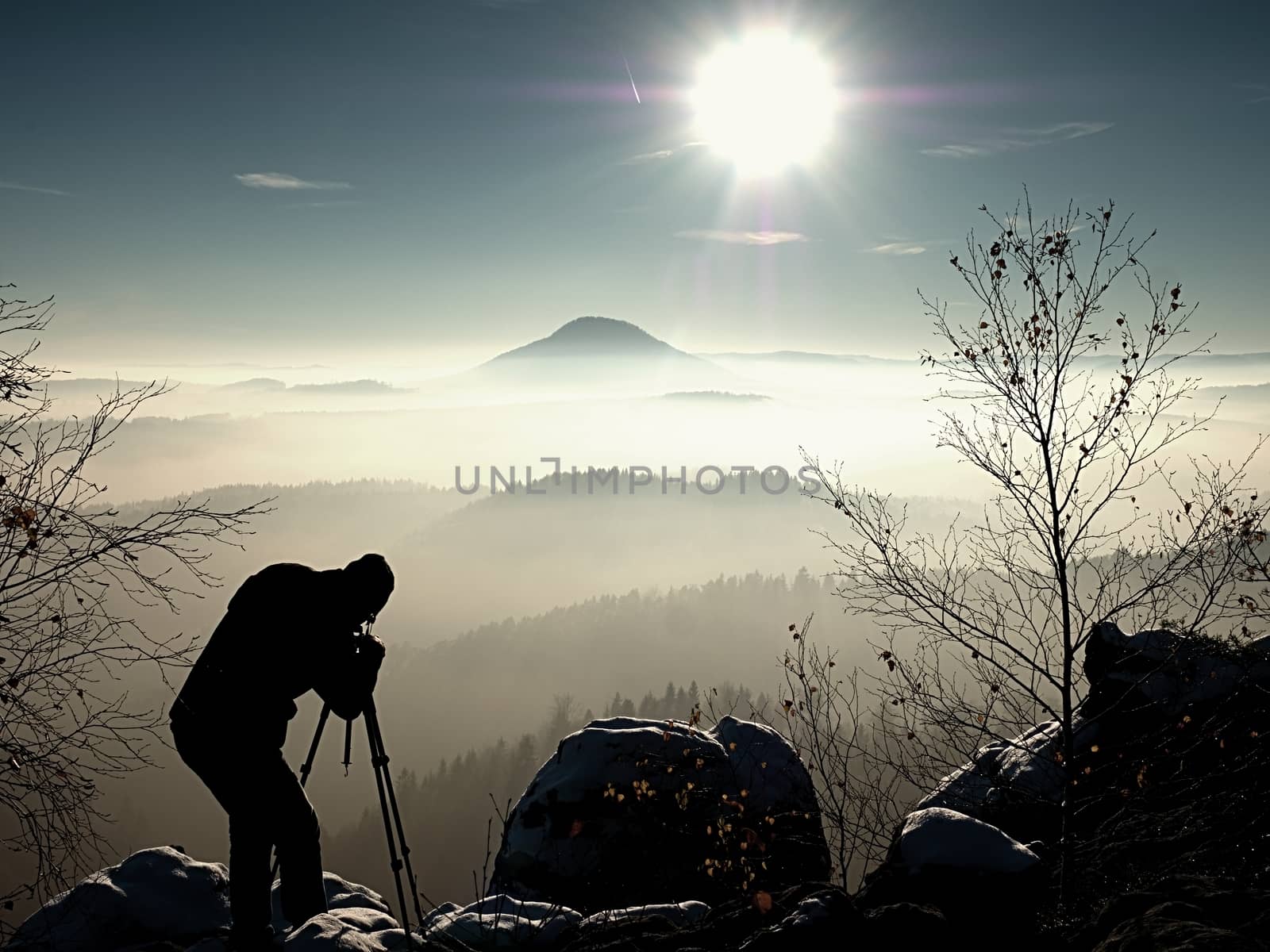 Sunny morning with first snow. Photographer preparing camera on tripod. Snowy rocks, in valley bellow colorful leaves forest. View over misty and foggy valley to Sun.