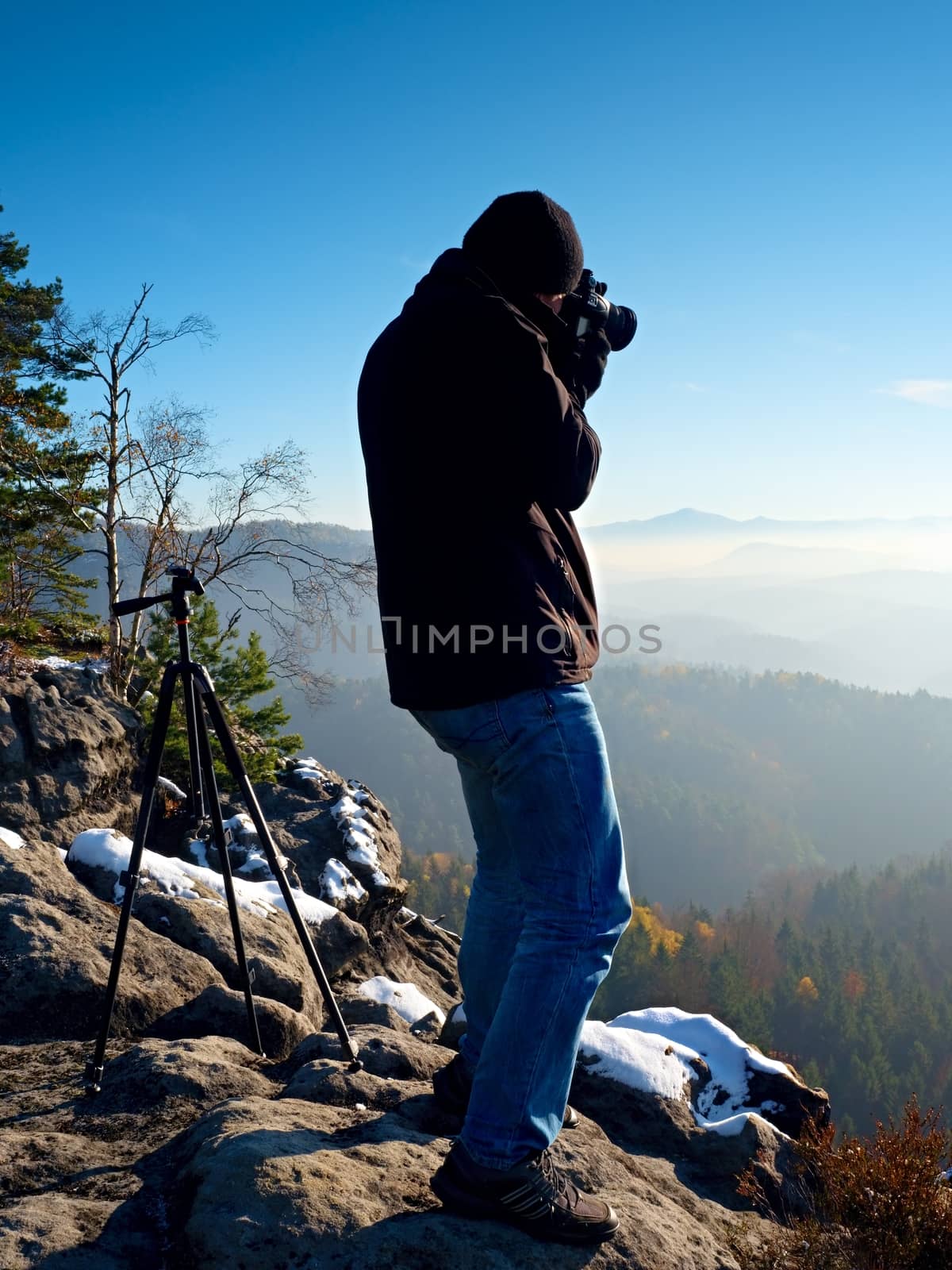 Professional nature photographer do work on snowy cliff.  Man takes photos with mirror camera on peak of rock. Dreamy fogy landscape, spring orange pink misty sunrise.