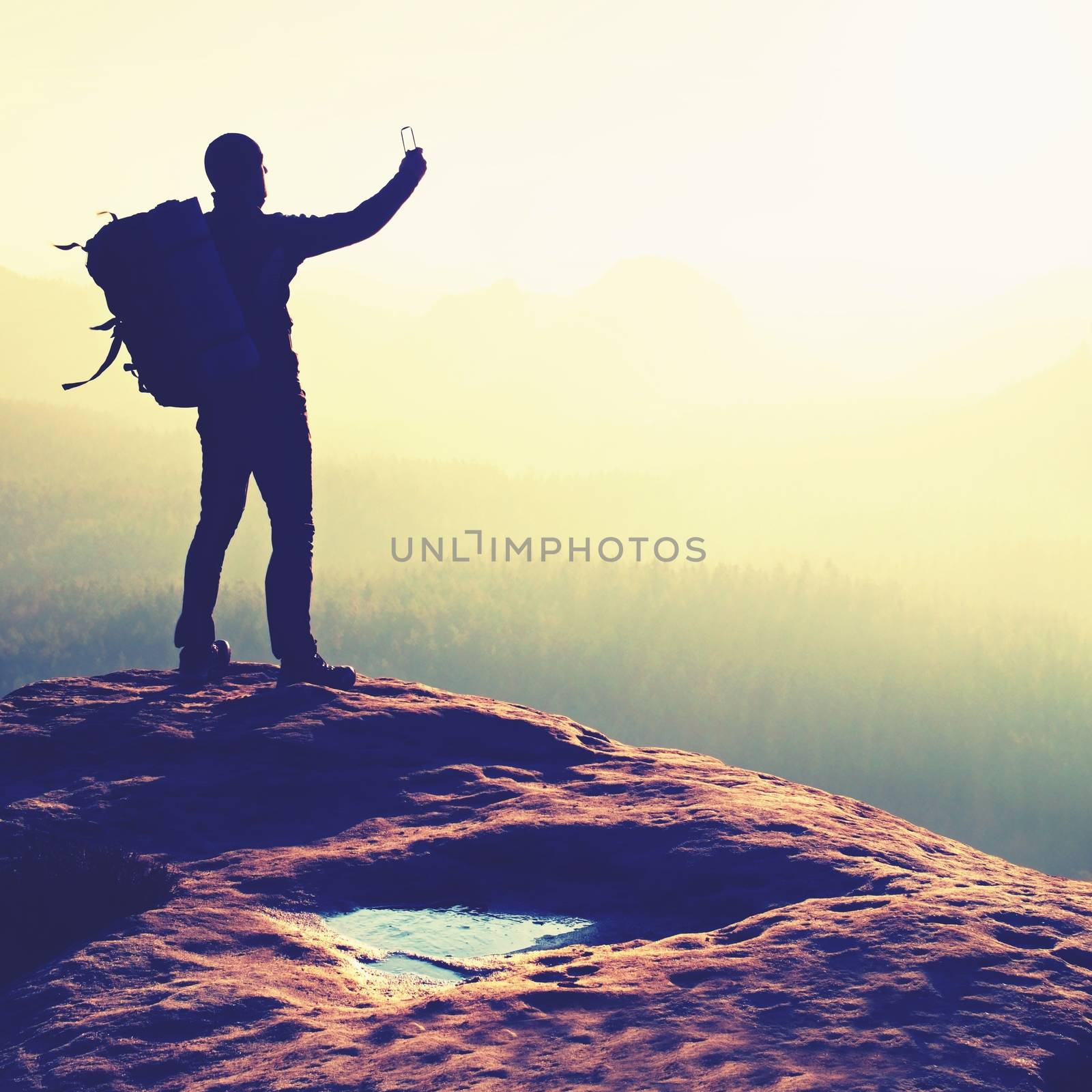 Tourist with backpack takes photos with smart phone on the rocky peak. Dreamy fogy valley below