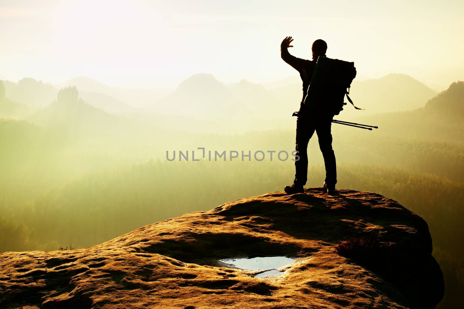 Tourist with backpack and poles in hand shadowing eyes. Sunny spring daybreak in rocky mountains. Hiker on rocky view point above misty valley. 