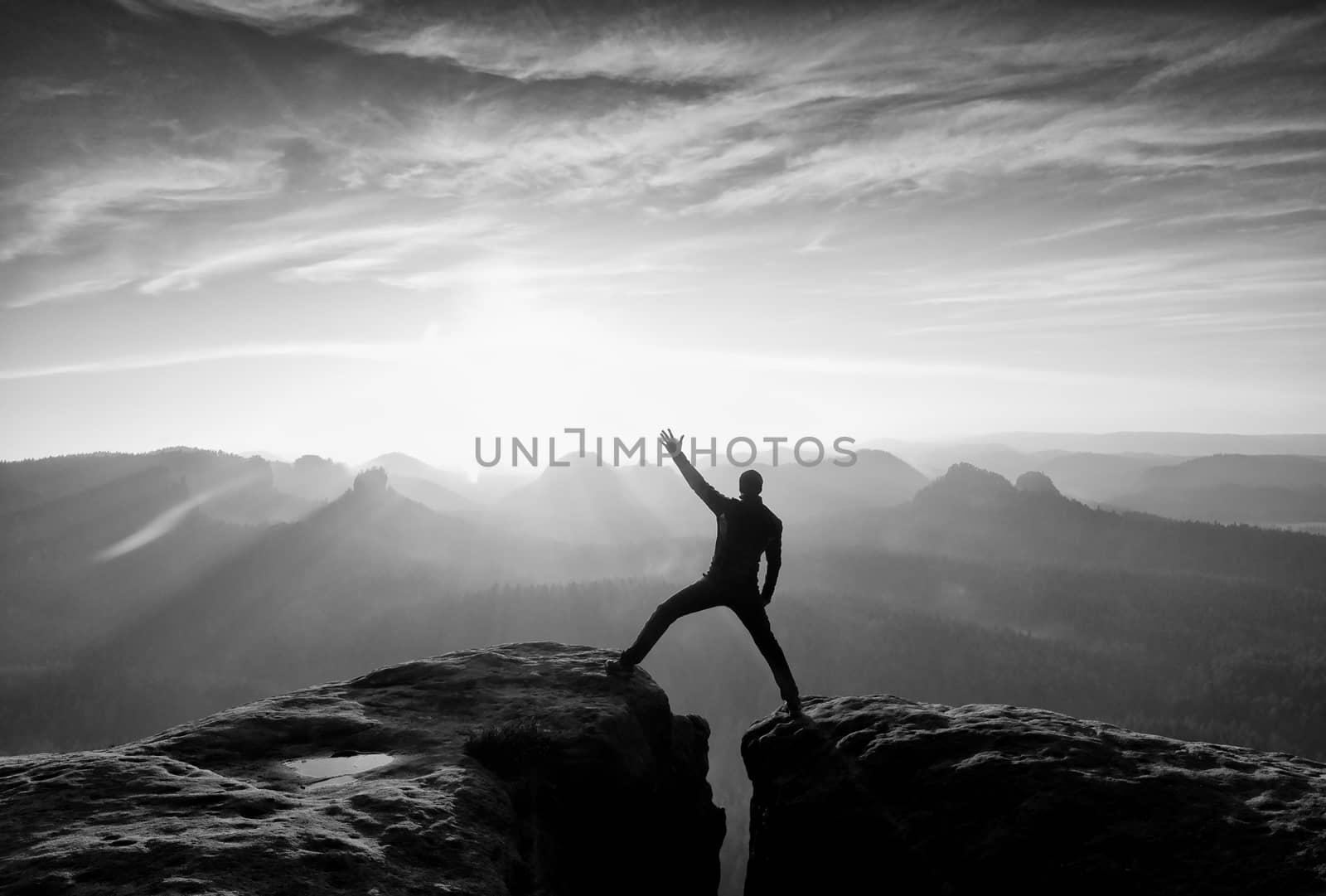 Jumping hiker in black gesture triumph between two rocky peaks. Wonderful daybreak in misty mountains.