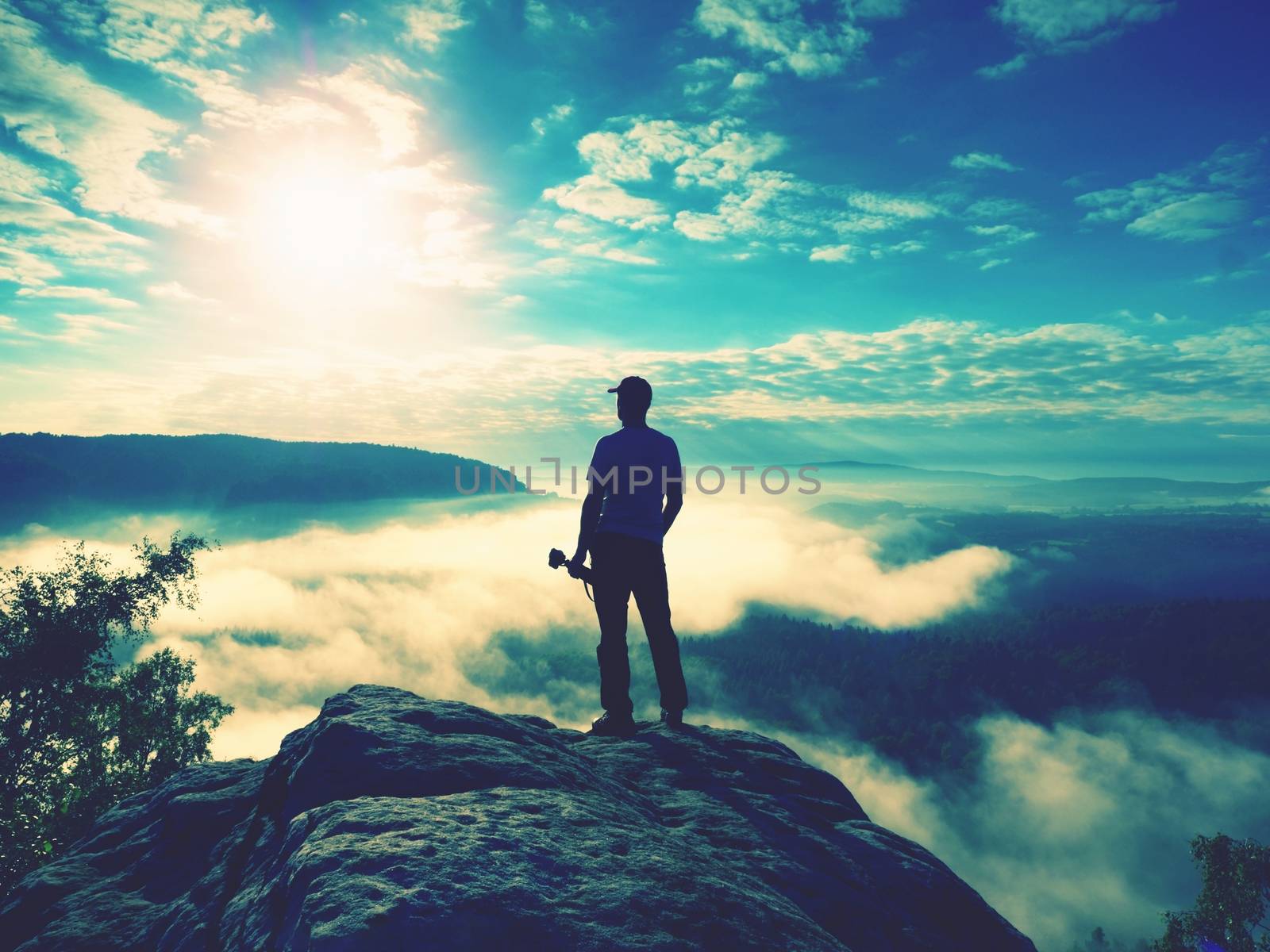 Tall photographer with baseball cap and tripod with camera in hands stand on rocky view point and watching down to deep misty valley bellow