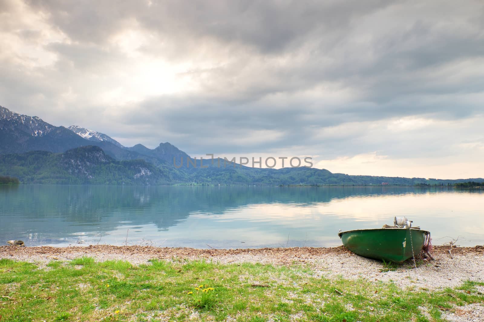 Abandoned fishing paddle boat on bank of Alps lake. Morning lake glowing by sunlight. Dramatic and picturesque scene. Mountains in water mirror.