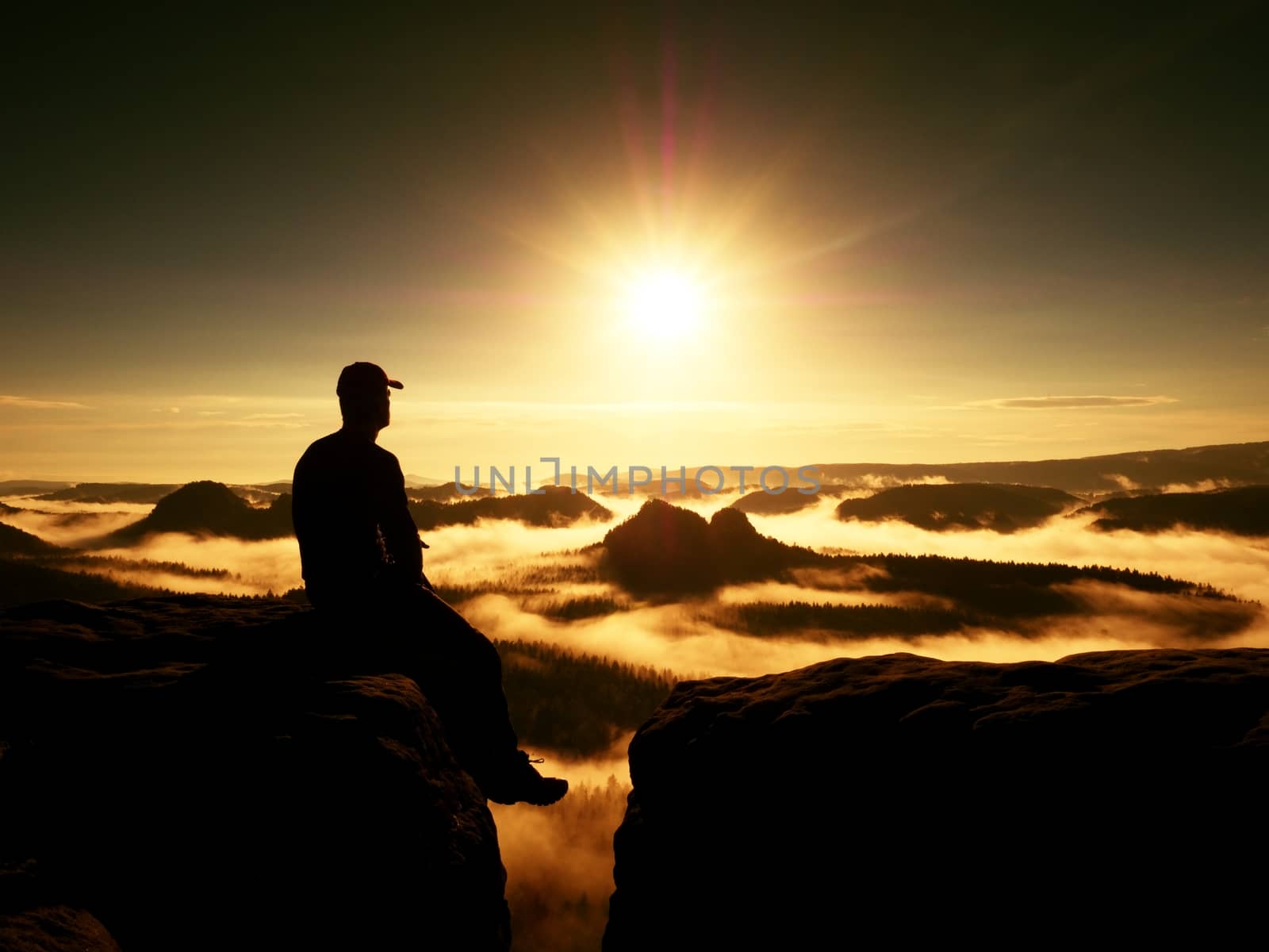 Moment of loneliness. Man with cap sit on the peak of rock and watching into colorful mist and fog in forest valley. Dreamy autumn ladscape