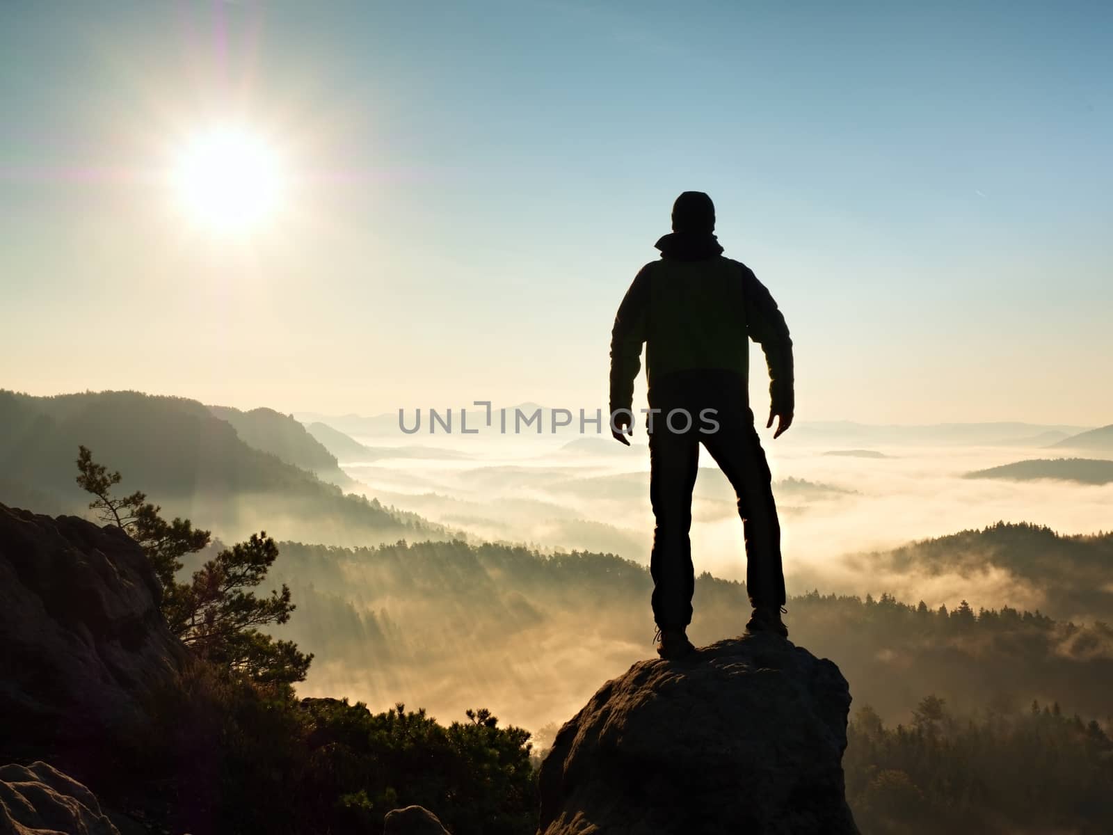 Autumnal  misty morning in nature. Hiker in black stand on peak in rock  and watching over the misty and foggy morning valley to Sun. 

