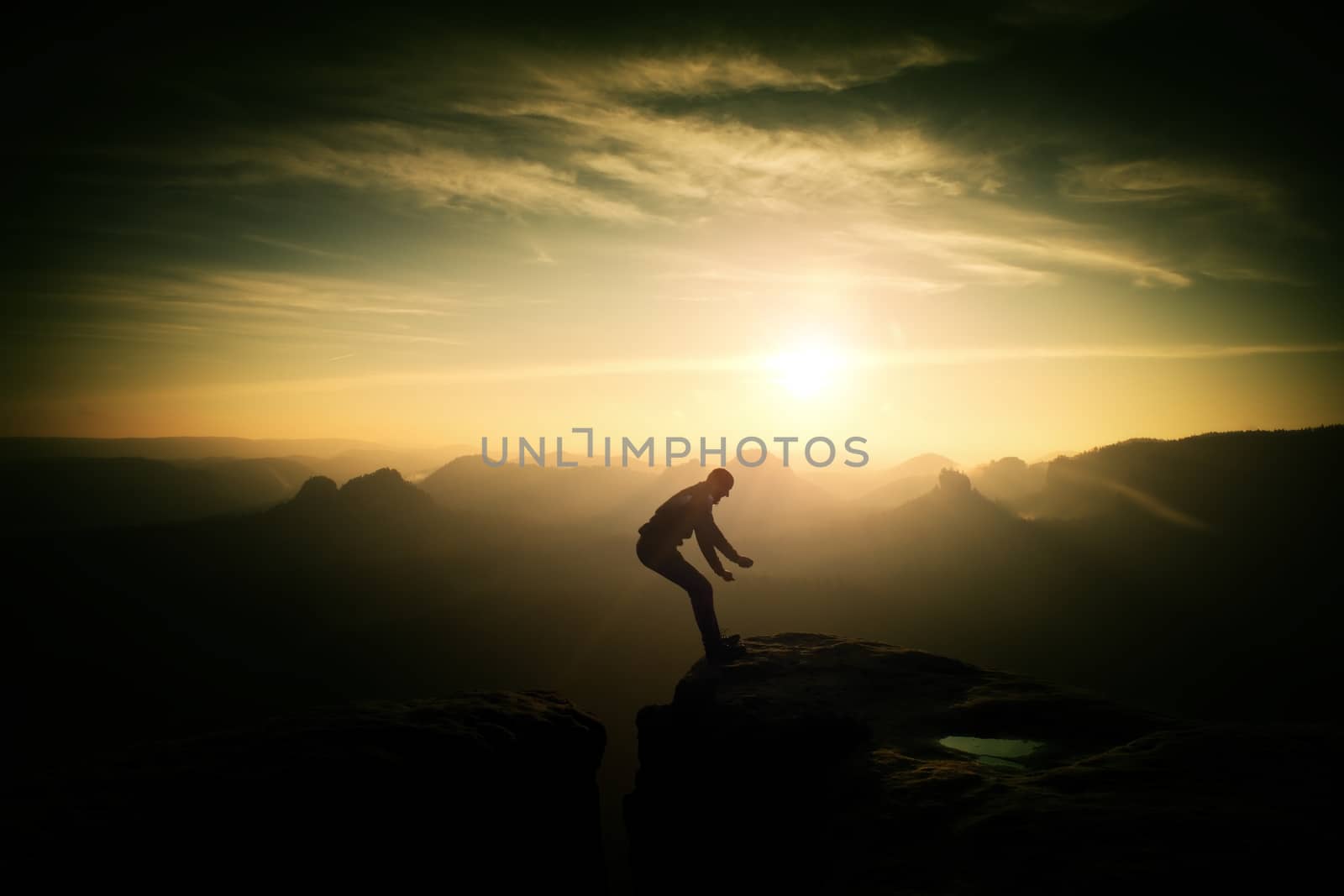 Crazy hiker umping between rocks. Marvelous colorful daybreak in rocky mountains, heavy orange mist in deep valley. Miracle of nature