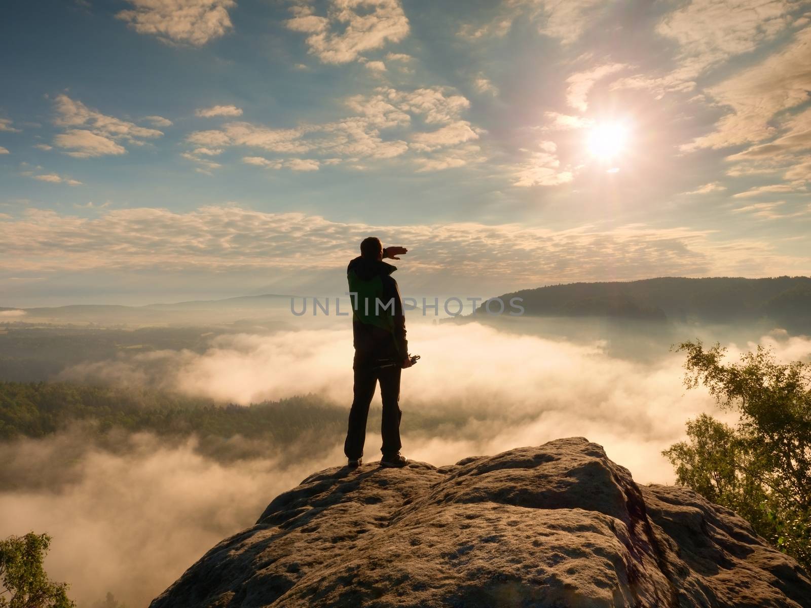 Professional photographer with tripod in hands on cliff and thinking. Dreamy fogy landscape, blue misty sunrise in a beautiful valley below