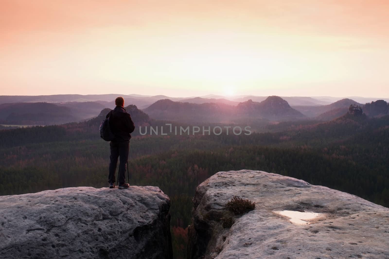 Hiker with sporty backpack stand on rocky view point above misty valley.  Sunny spring daybreak in rocky mountains.