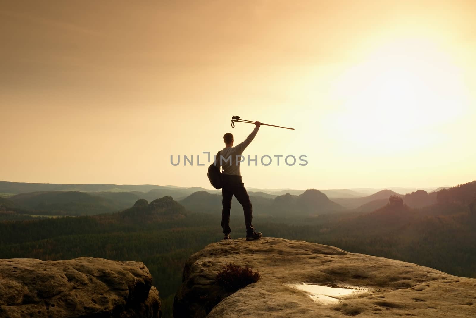 Hiker on peak. Man in his target with trekking poles in hand in air. Hiker in black running trousers and shinning blue sweaty t-shirt,