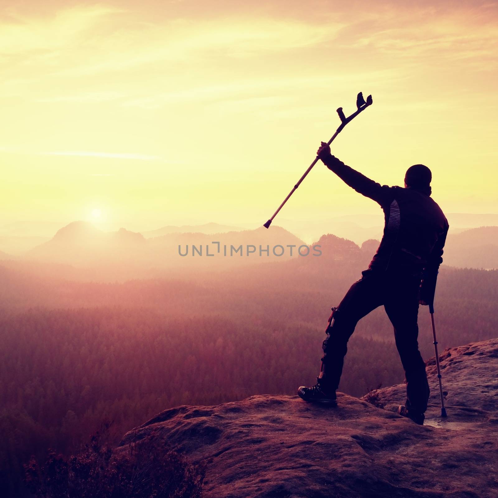 Hiker with  medicine crutch above head achieved mountain peak. Broken leg fixed in immobilizer Deep misty valley bellow silhouette of man with hand in air. Spring daybreak