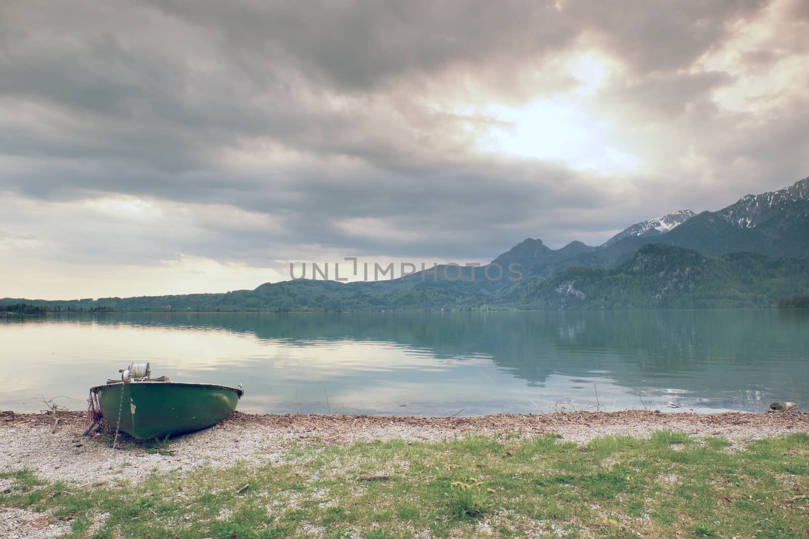 Abandoned fishing paddle boat on bank of Alps lake. Morning lake glowing by sunlight. Dramatic and picturesque scene. Mountains in water mirror.