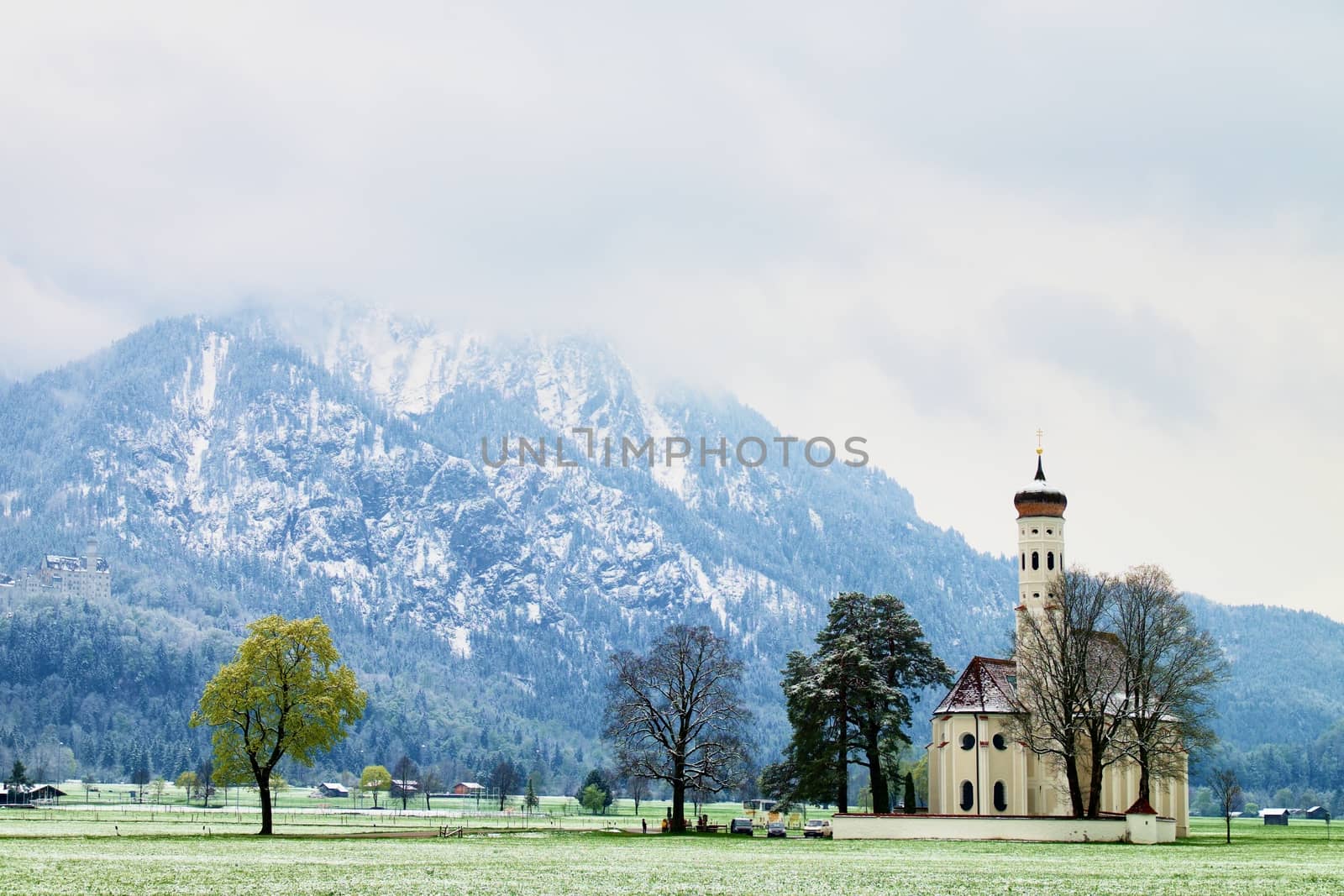 Old church with high bell tower and graveyard bellow mountains.  April weather. Wet spring snow in already fresh green grass  of meadow bellow mountains. 