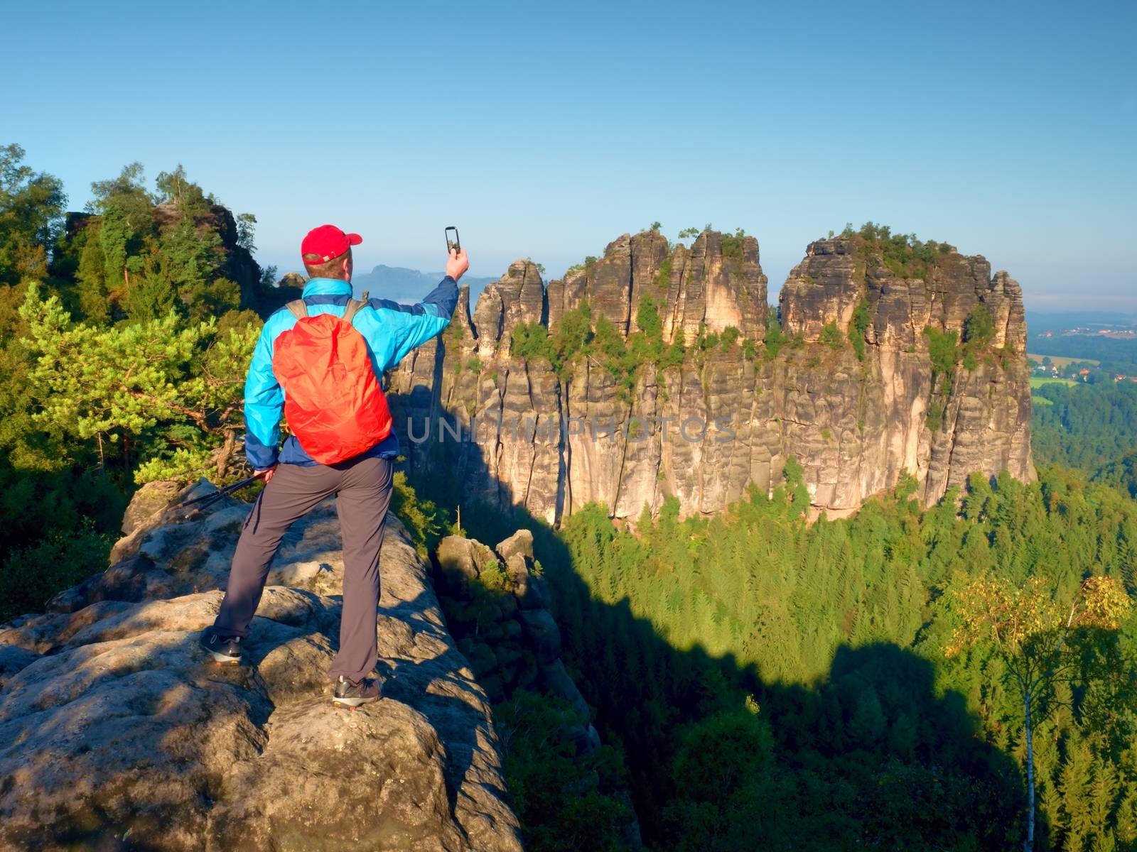 Tourist with red backpack takes photos with smart phone of rocks and valley. Spring or summer sunny weather, fresh green colors