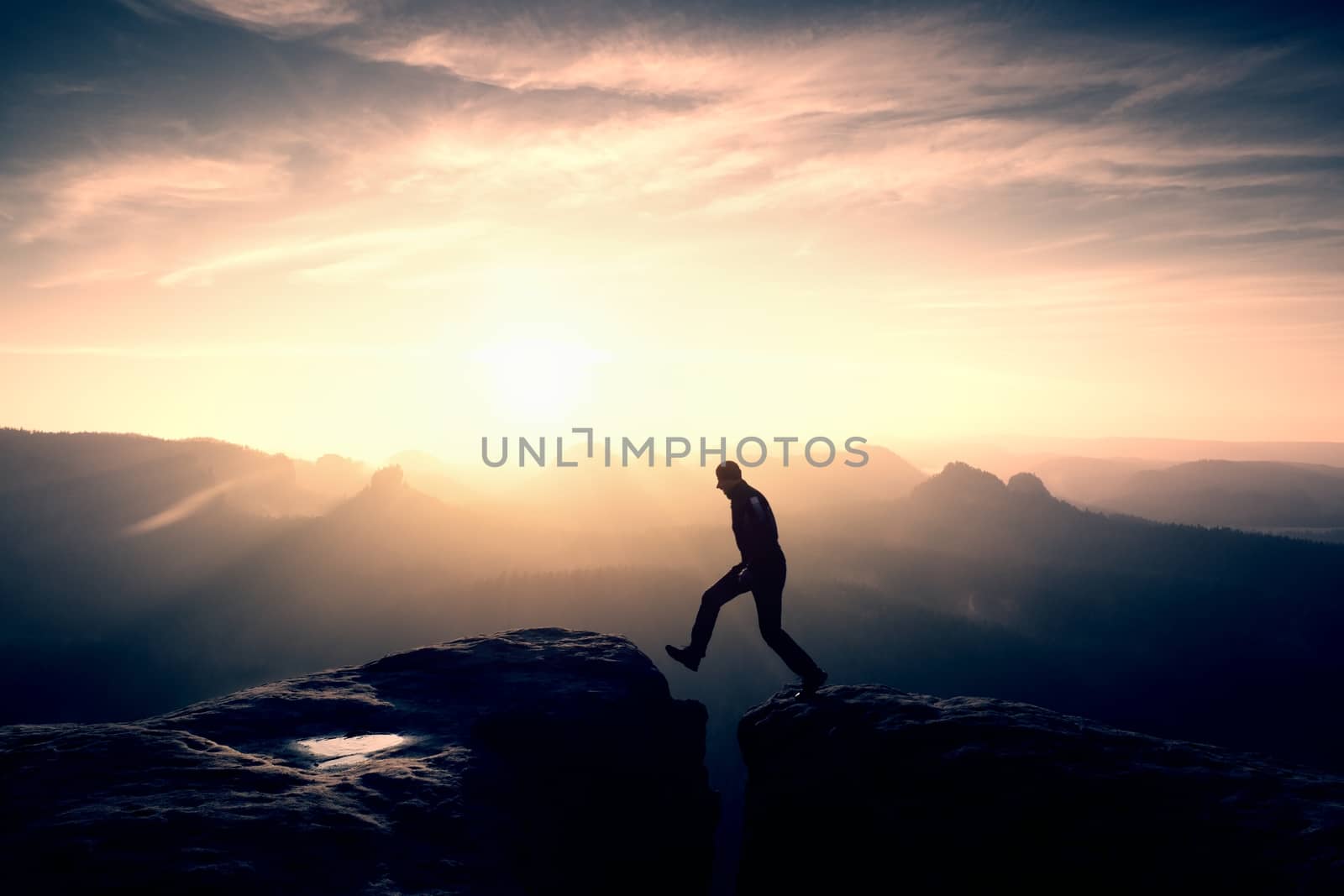 Crazy hiker umping between rocks. Marvelous colorful daybreak in rocky mountains, heavy orange mist in deep valley. Miracle of nature