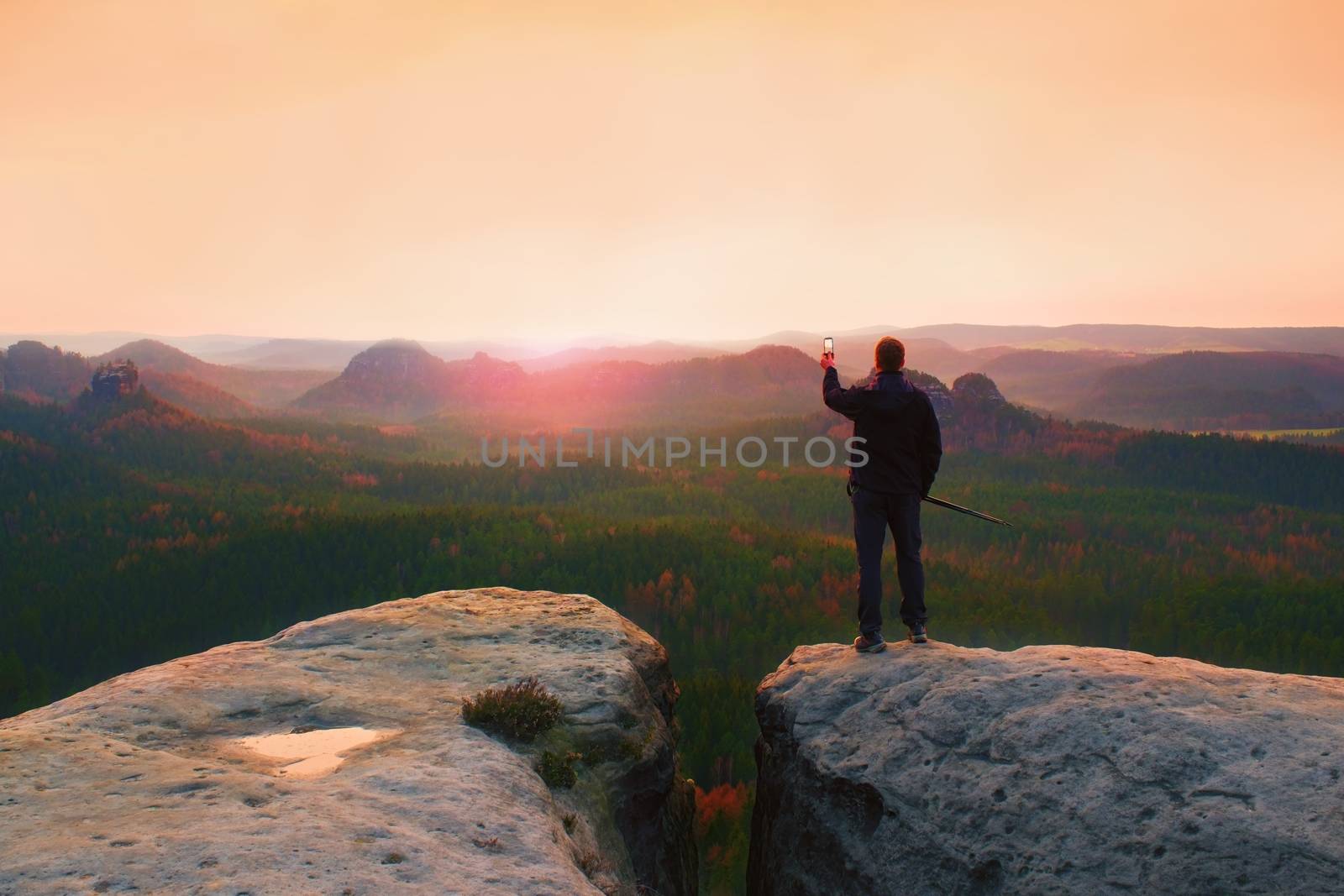 Hiker stand with smart phone on top of mountain and enjoying spring sunrise. Tourist guide with trekking poles.