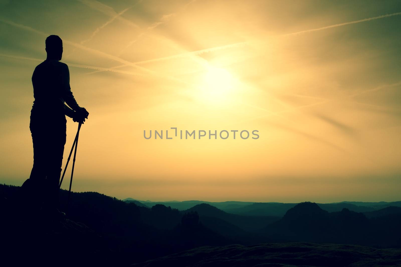 Silhouette of tourist with poles in hand. Sunny spring daybreak in rocky mountains. Hiker w on rocky view point above misty valley. 