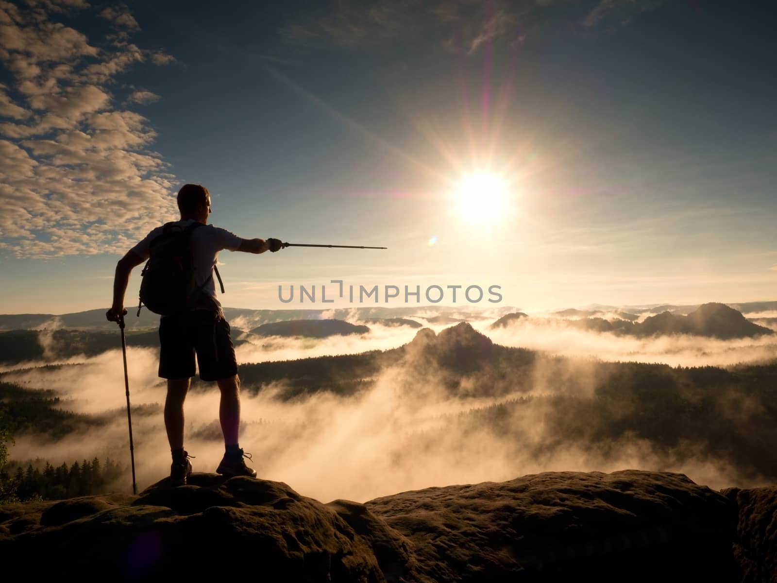 Tall backpacker with poles in hand. Sunny misty daybreak in rocky mountains. Hiker with backpack stand on rocky view point above valley. Vivid and strong vignetting effect.