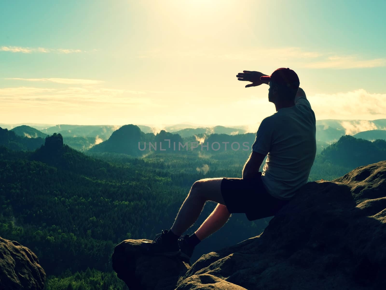 Man sit on the peak of rock. Hiker shadowing eyes with raised arm,  watching into colorful mist and fog in forest valley.