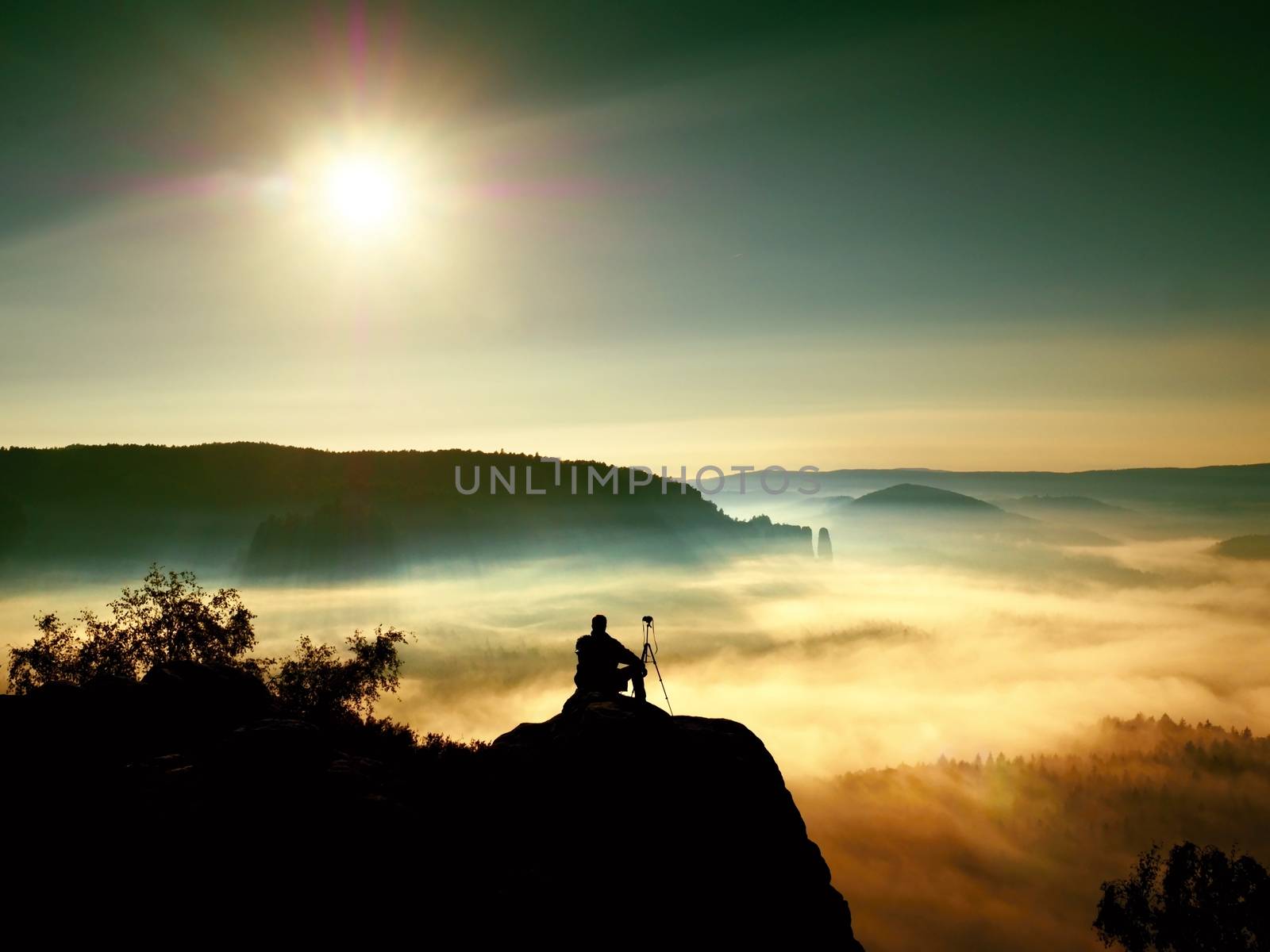 Nature photographer with tripod and camera on cliff and thinking. Dreamy fogy landscape, orange misty sunrise in a beautiful mountains