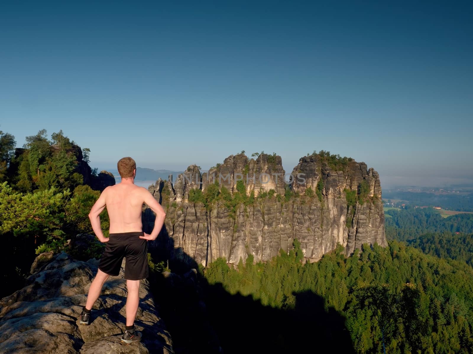 Naked man in black pants only at the top of the rock side.  Sharp sandstone cliffs above deep valley. Popular climbers resort.