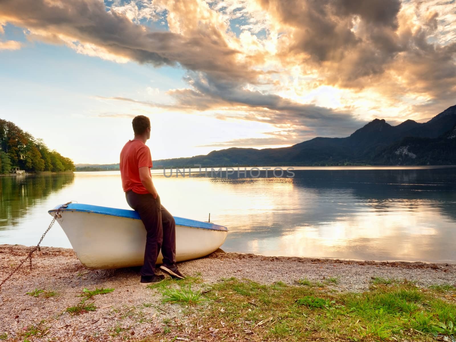 Tiired man in red shirt sit on old fishing paddle boat at mountains lake coast. Afternooon sun hidden in clouds above mountain peaks. Vintage photo effect