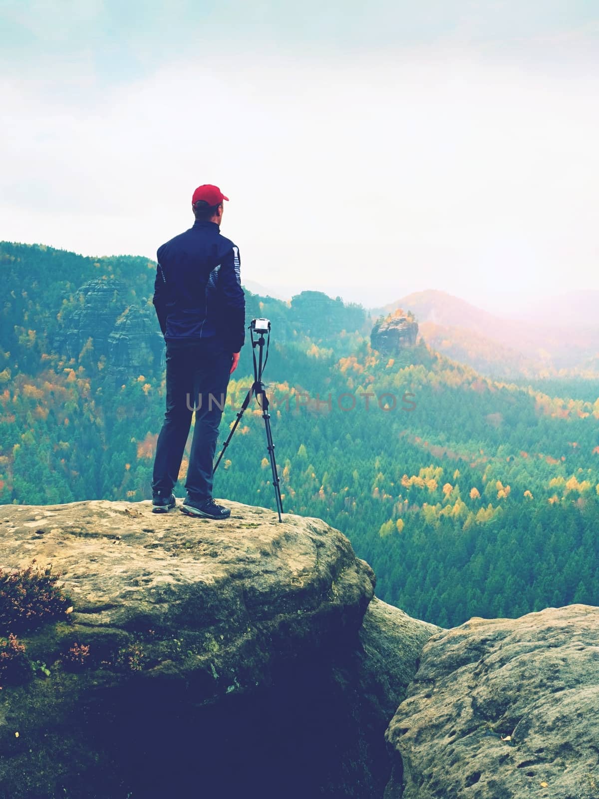 Outdoor photographer with tripod and camera on rock thinking. Creamy mist in autumnal valley bellow. Dreamy rocky mountains. Misty sunrise in a beautiful valley 