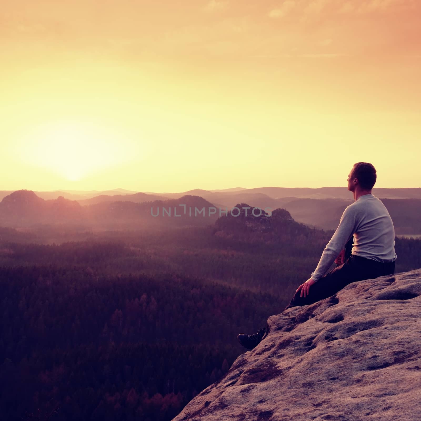 Man sit on the peak of rock and watching into colorful mist and fog in forest valley. Hiker enjoying view of morning nature