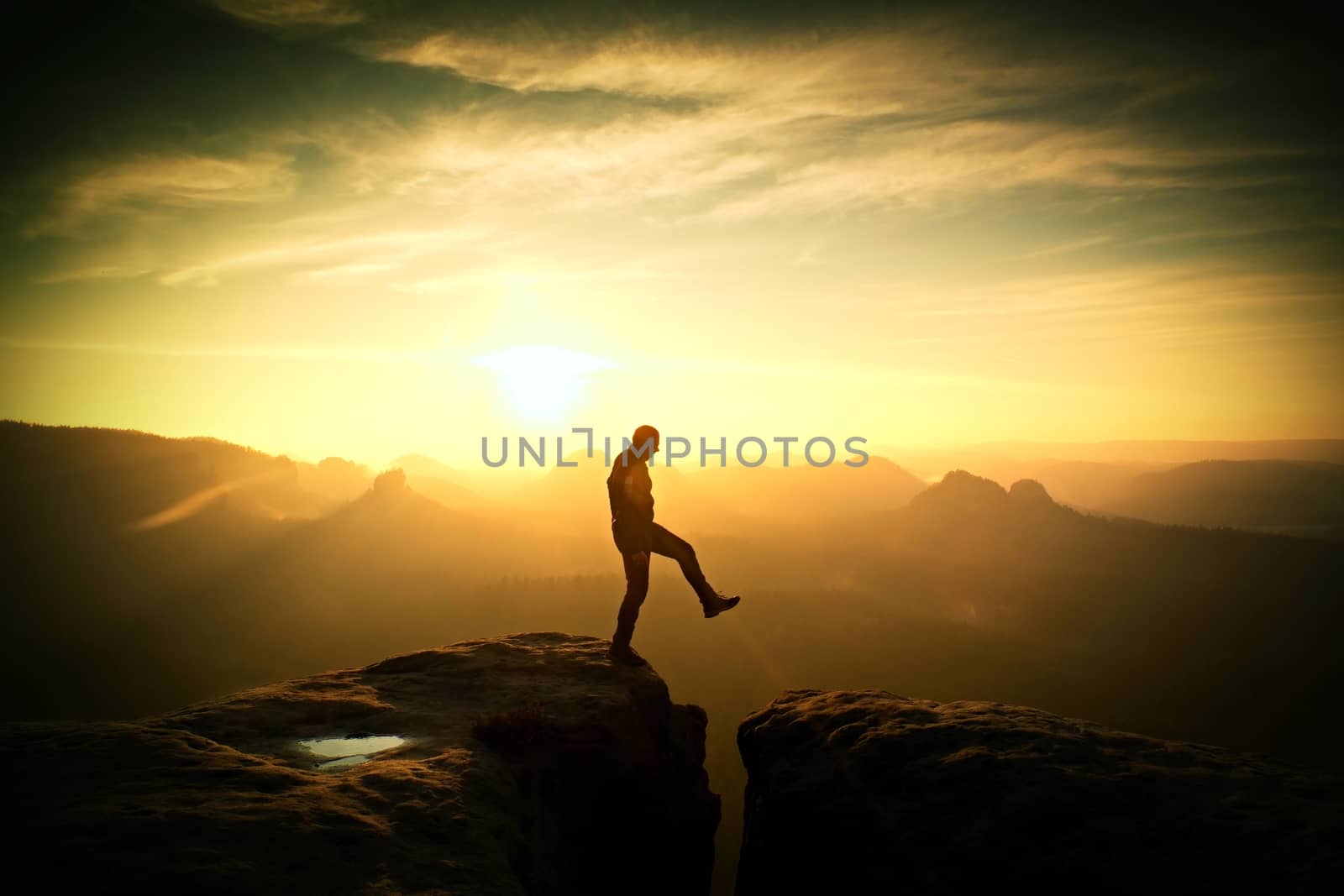 Crazy hiker umping between rocks. Marvelous colorful daybreak in rocky mountains, heavy orange mist in deep valley. Miracle of nature